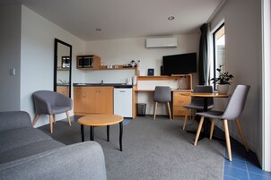 Una sala de estar moderna con un sofá, una mesita redonda y un escritorio con dos sillas. Hay una zona de cocina con microondas, fregadero y refrigerador. Un televisor de pantalla plana está montado en la pared, y hay una planta en maceta sobre el escritorio.