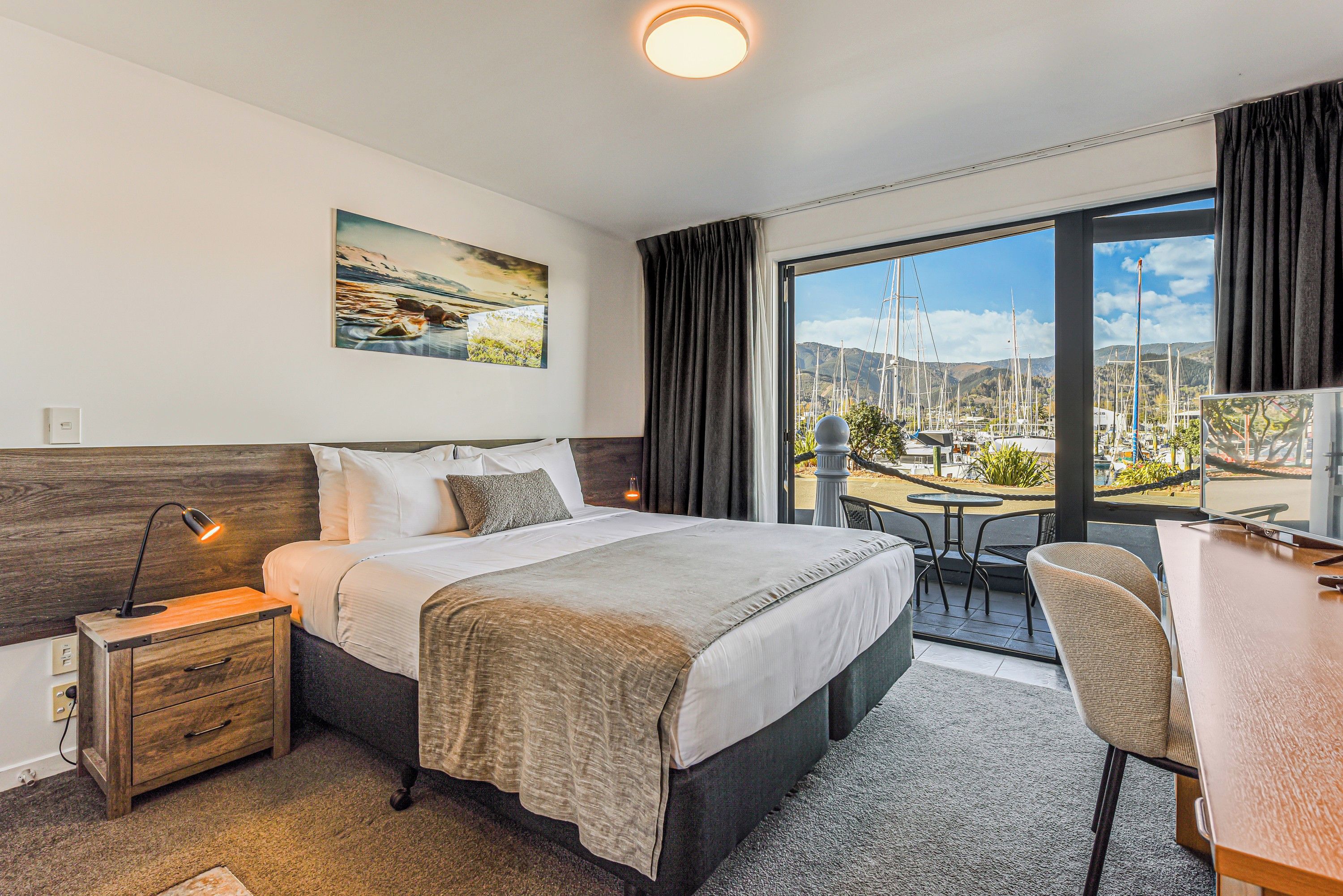 A hotel room with a bed, bedside table, desk, and a view of a marina through a large window.