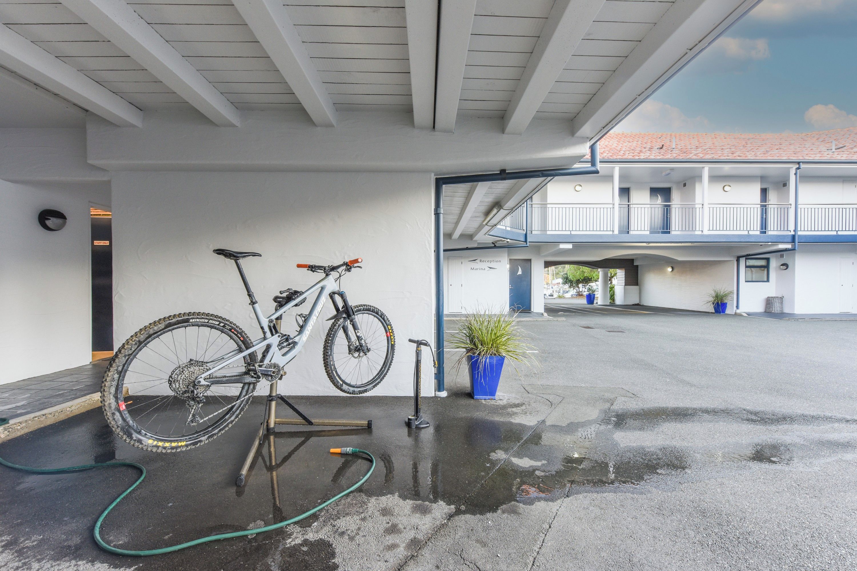 Uma bicicleta estacionada sob uma cobertura, com o chão molhado, uma scooter e um vaso de planta.