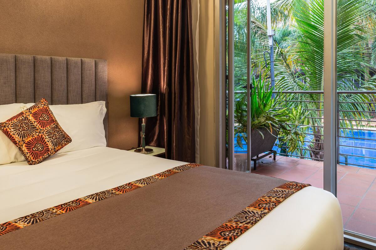 A hotel room with a bed, a nightstand, a lamp, and a view of a pool and greenery.
