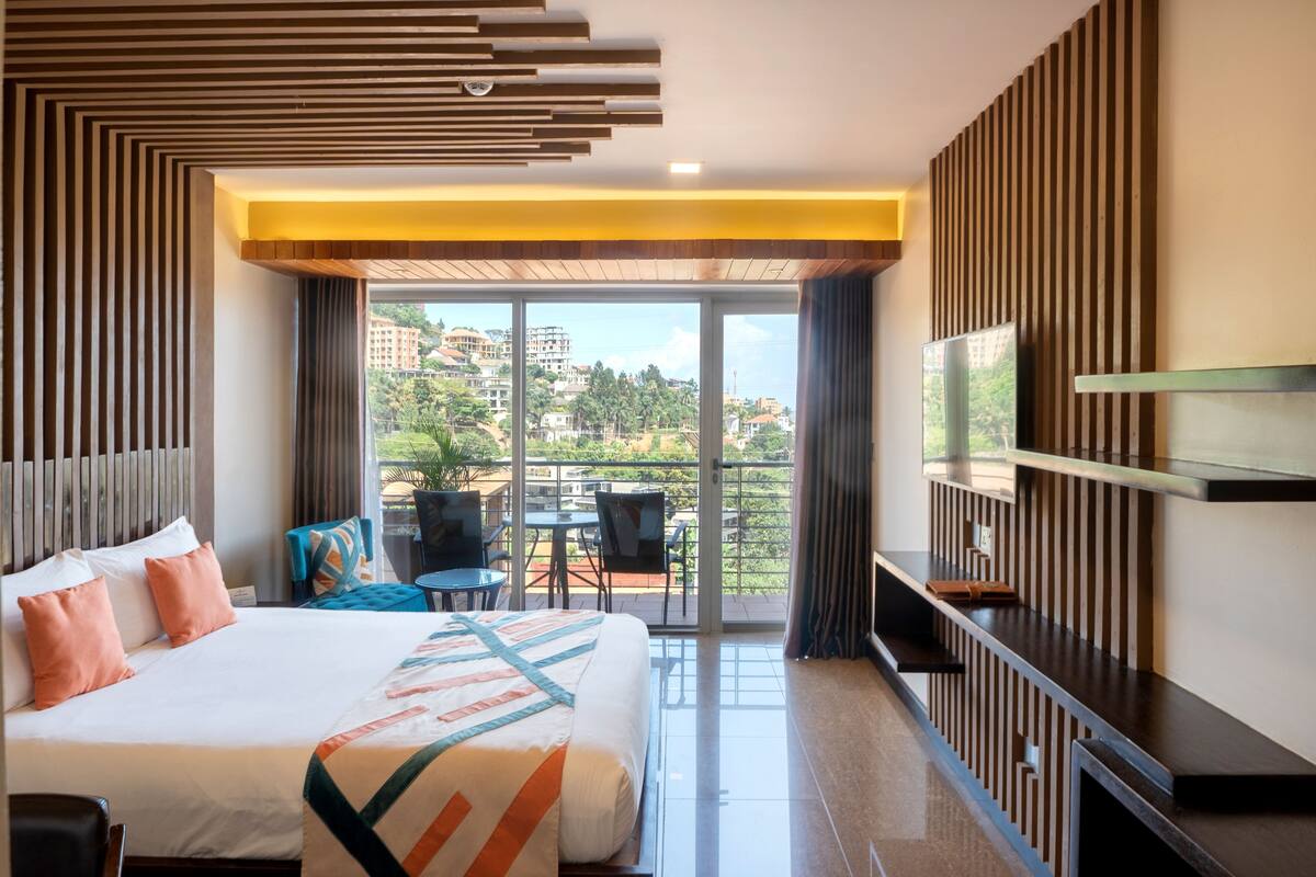 A modern hotel room with a large bed, a TV, a balcony with a view of buildings and greenery, and a seating area with chairs and a table.