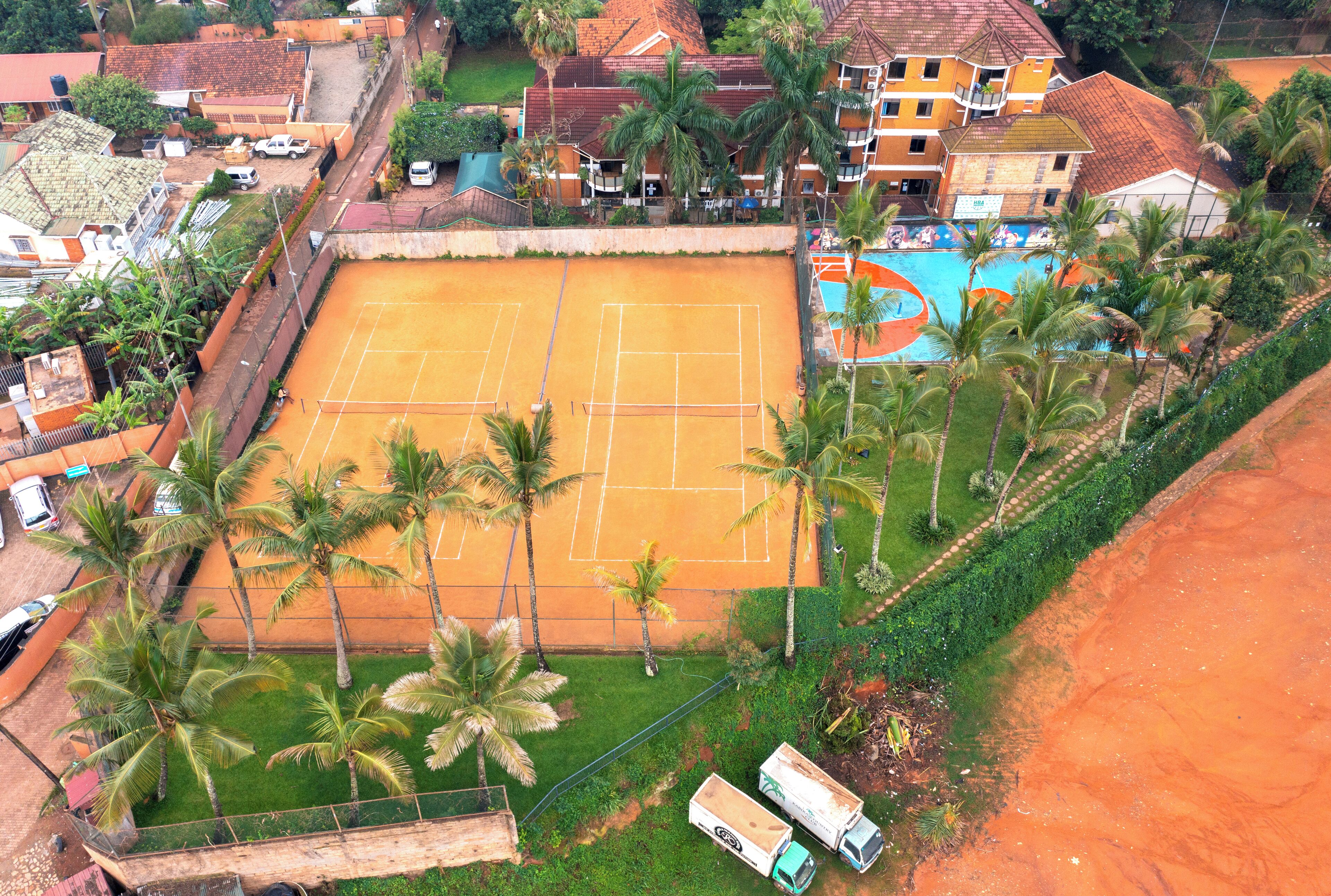 A tennis court surrounded by palm trees, a pool, and a building with a red roof.