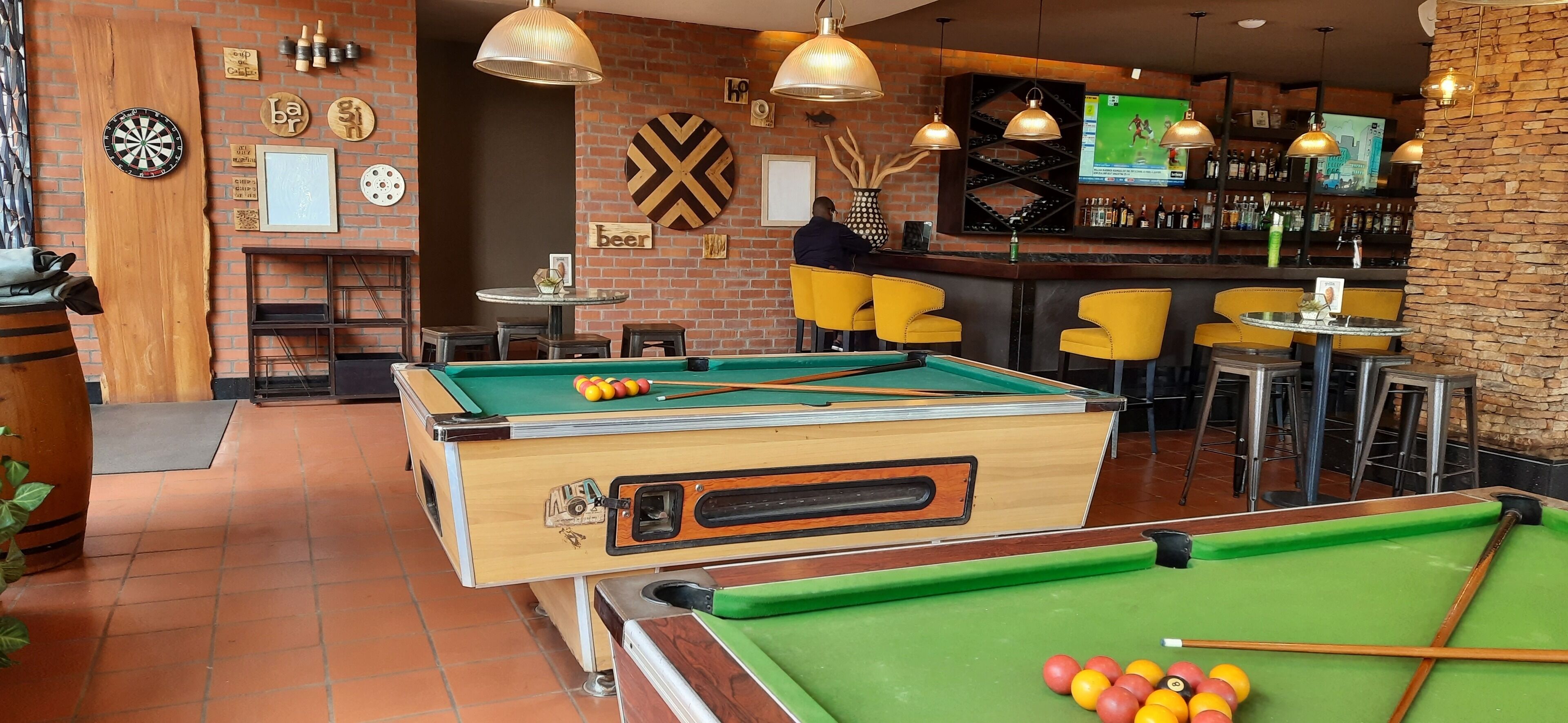 Una sala de billar con mesa de pool, dardos y zona de bar con sillas amarillas y una pantalla de televisión.