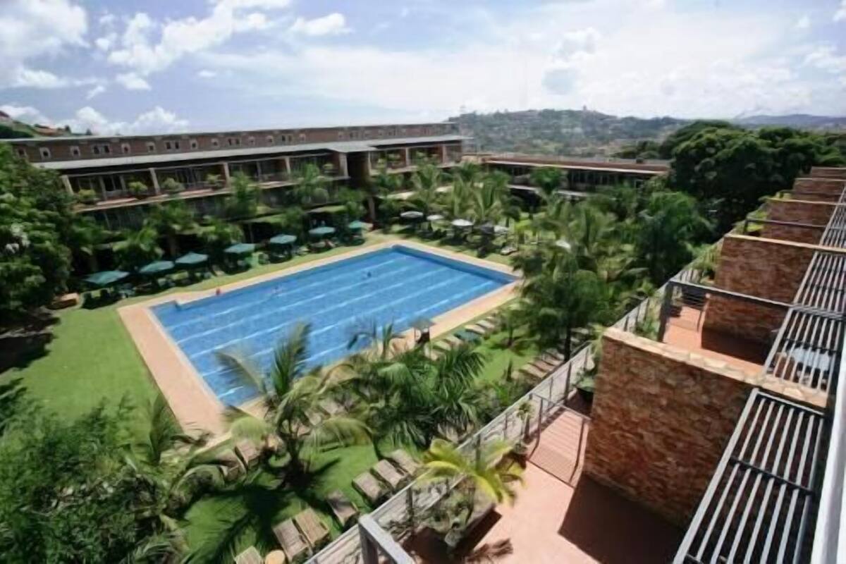 A swimming pool surrounded by greenery, a building with multiple balconies, and a mountainous backdrop.