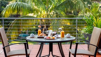 A round table with breakfast items, two wicker chairs, and a balcony with a metal railing and tropical plants.