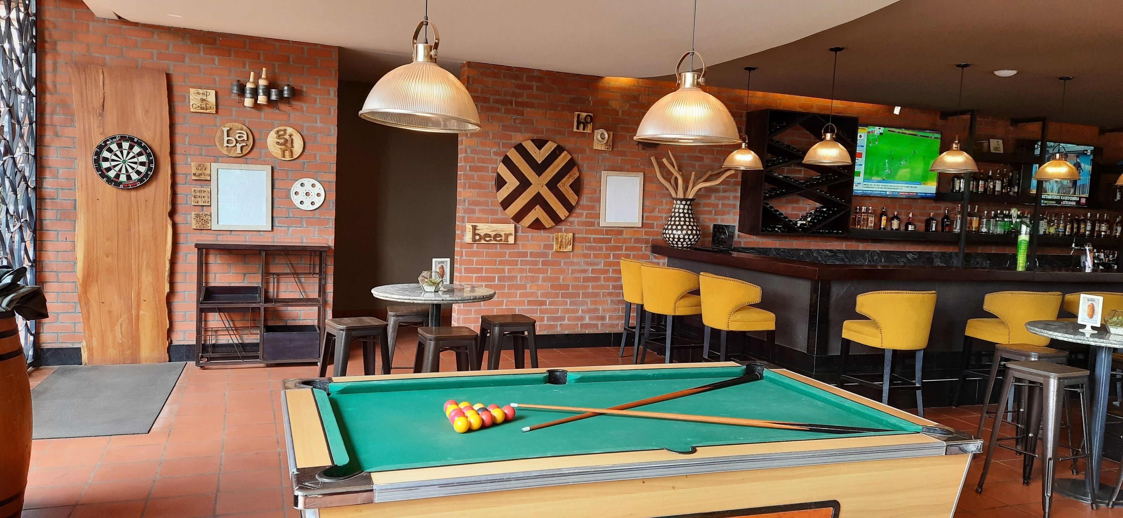 Mesa de billar con bolas y tacos, un dardo y una zona de bar con sillas amarillas y una pantalla de televisión.