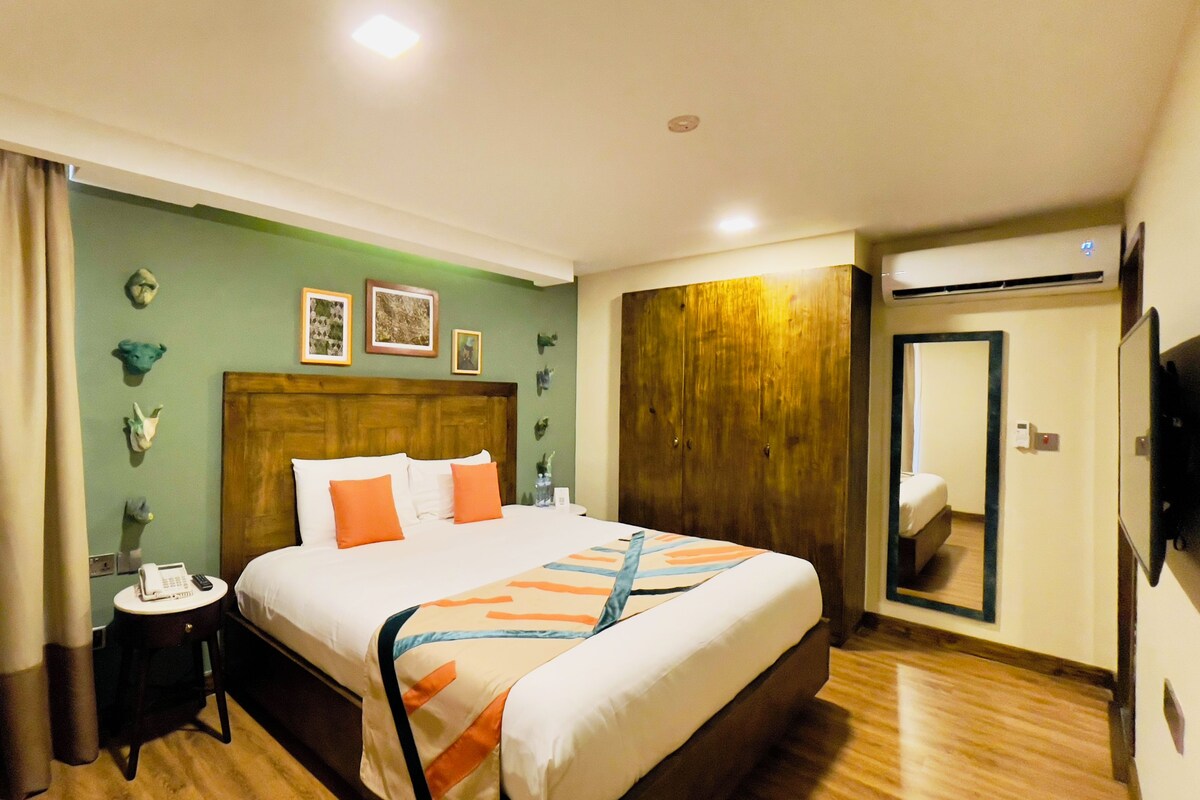 A hotel room with a large bed, a wooden wardrobe, a bedside table with a lamp, and a mirror on the wall.