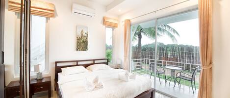 Un dormitorio con una cama grande, una mesita de noche, un espejo y una ventana con vista a un balcón y palmeras.