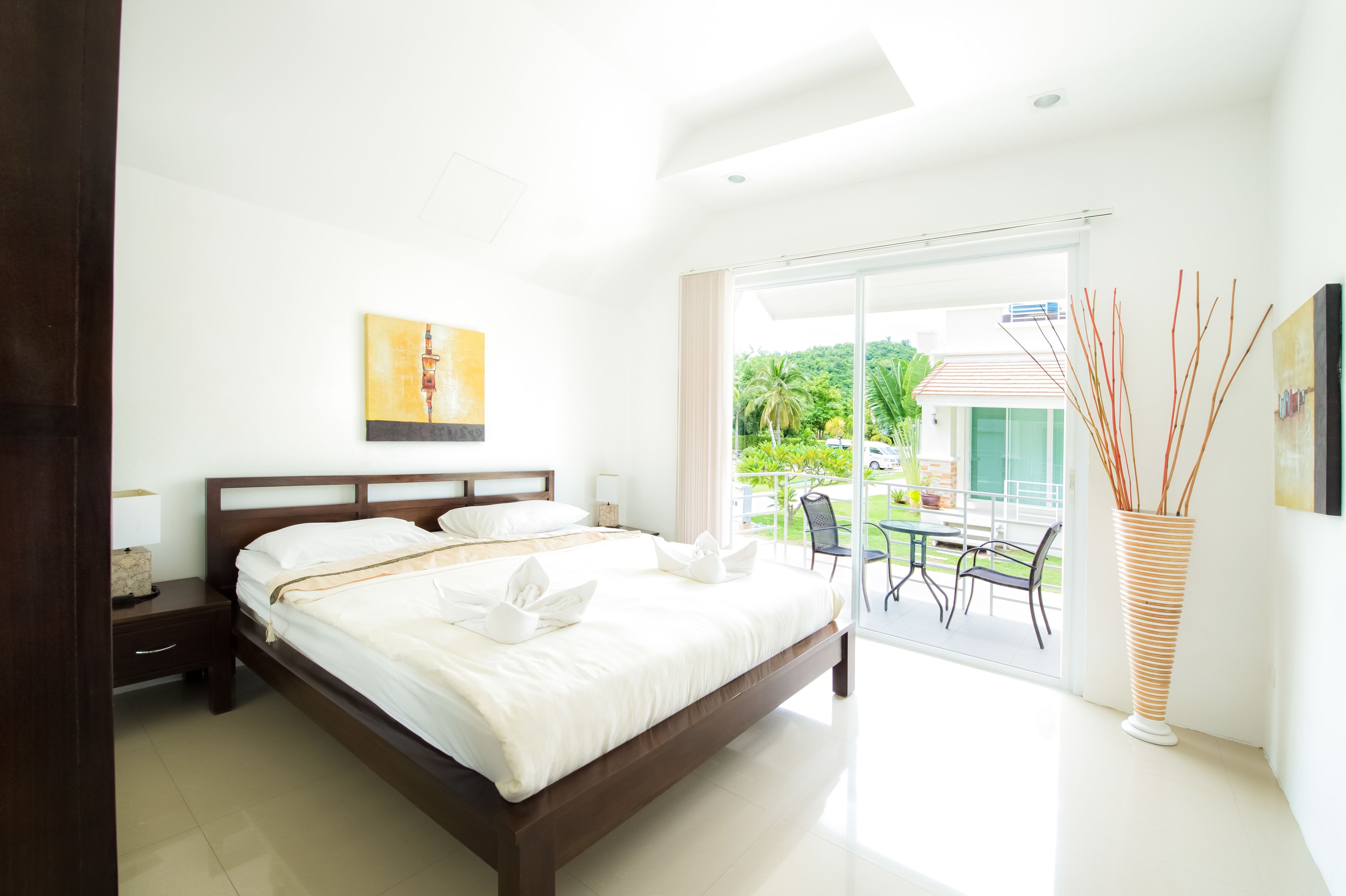 A modern hotel room with a large bed, a nightstand, a bedside table, a window with a view of greenery, and a vase with decorative sticks.
