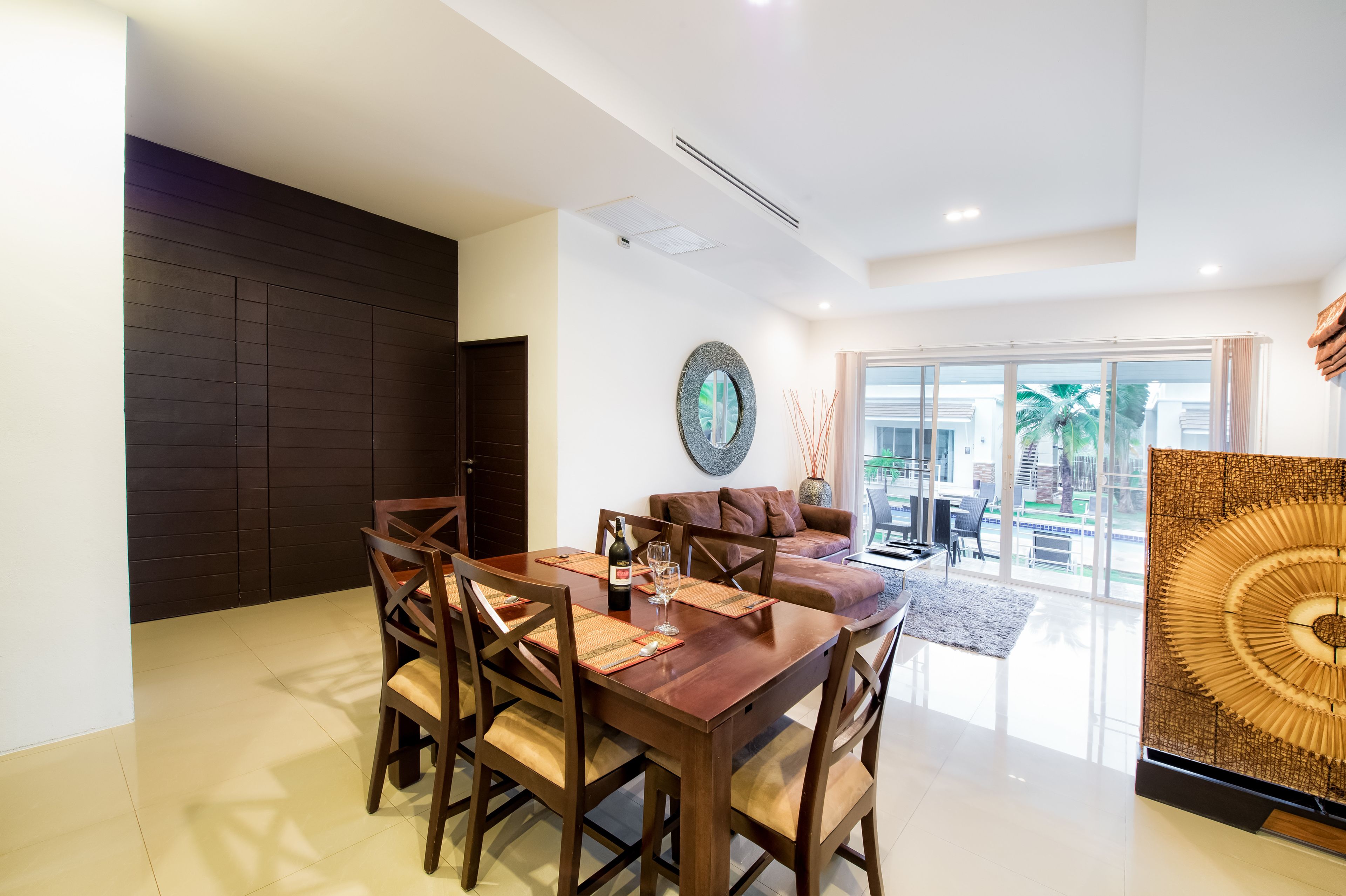 A modern dining area with a wooden table and chairs, a round mirror, and a view of the outdoors through large glass doors.