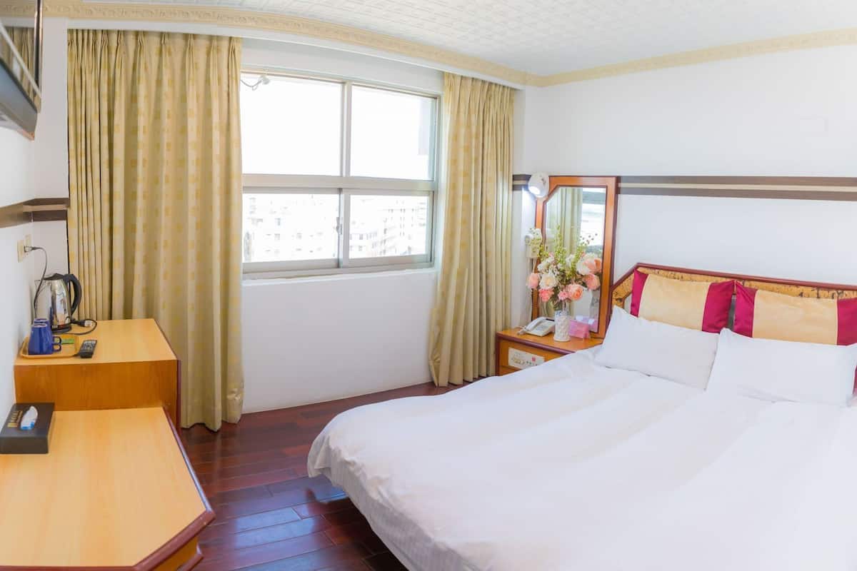 A hotel room with a bed, a desk, a mirror, a window with curtains, and a view of buildings outside.