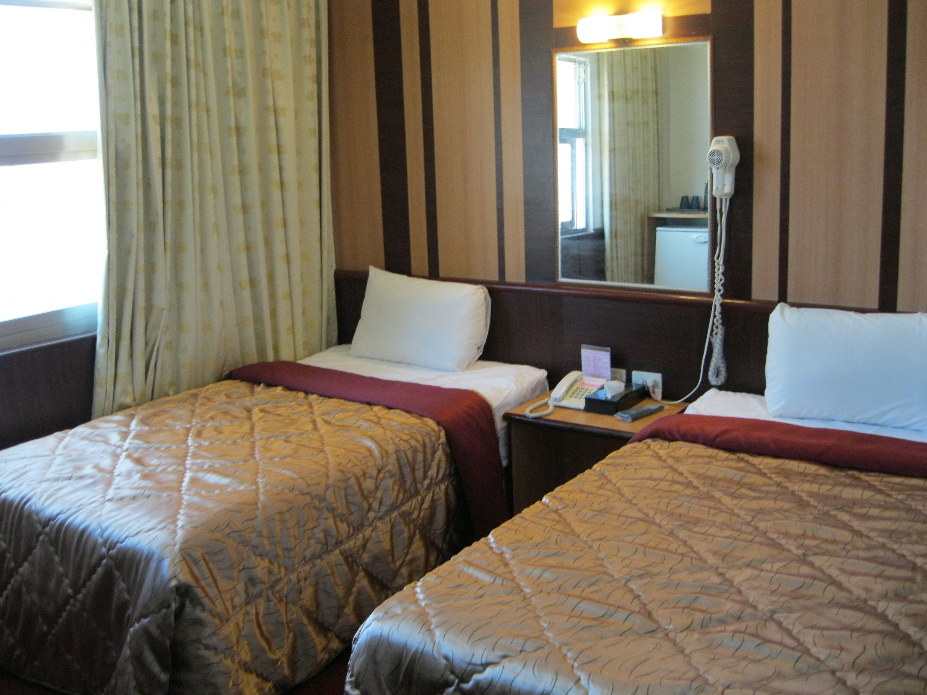 Une chambre d’hôtel avec deux lits, une table de chevet avec un téléphone et une lampe, et une fenêtre avec des rideaux.