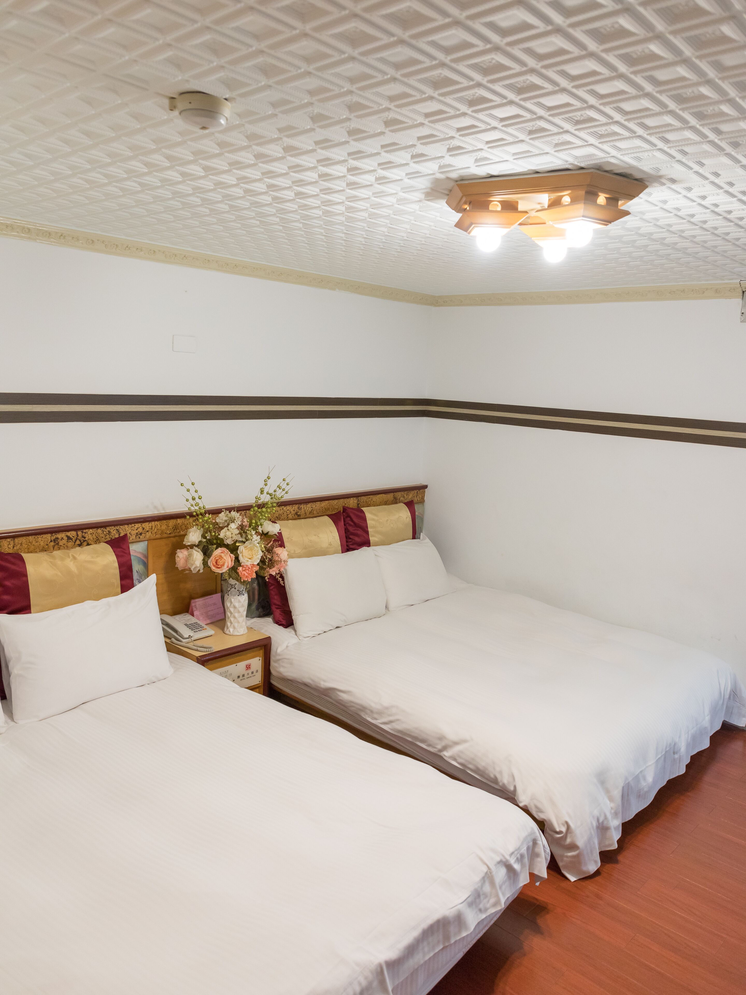 Habitación de hotel con dos camas, un cabecero de madera, una mesita de noche con un teléfono y un jarrón con flores.