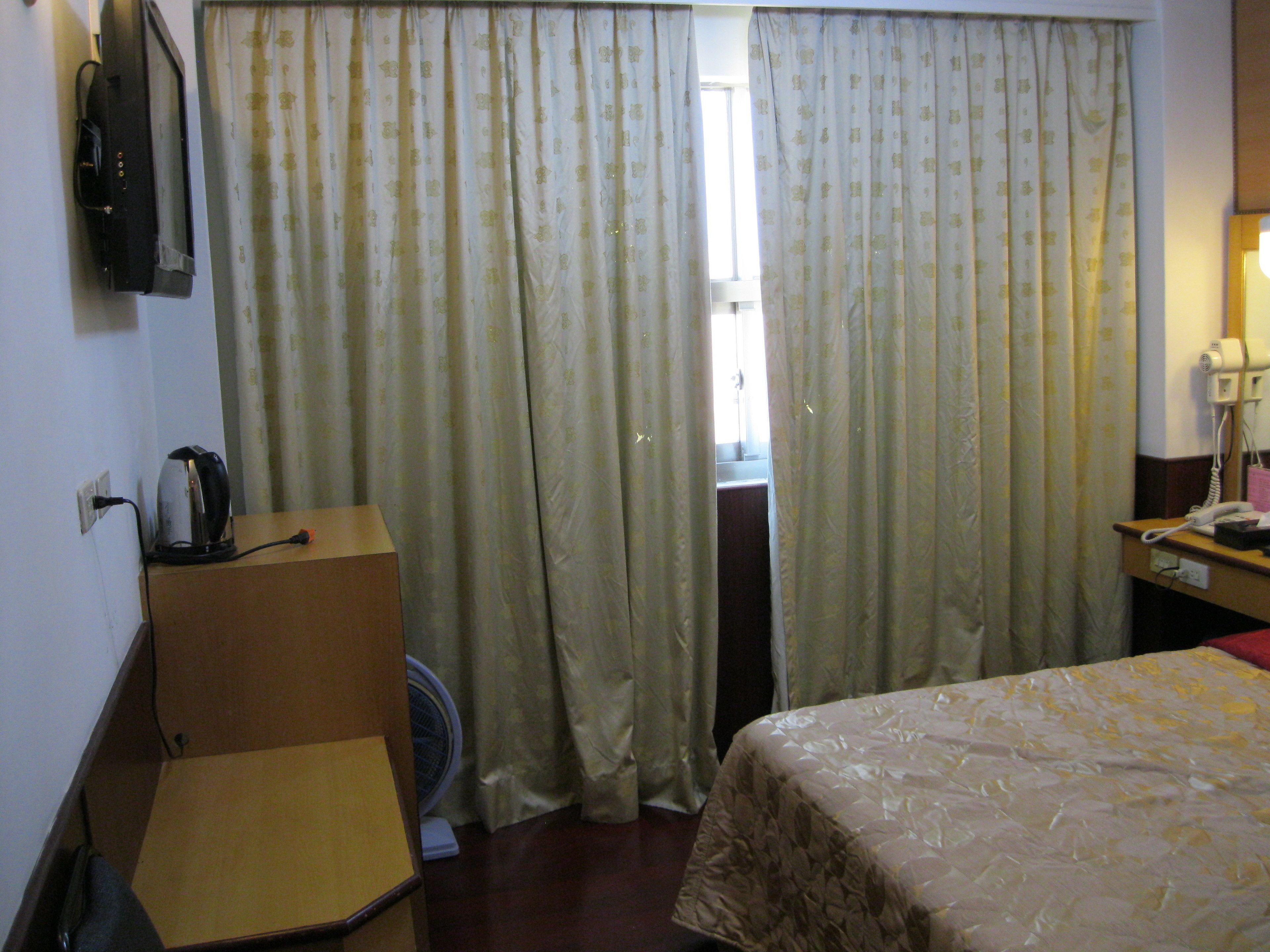 A hotel room with a bed, a desk, a kettle, a fan, and curtains.