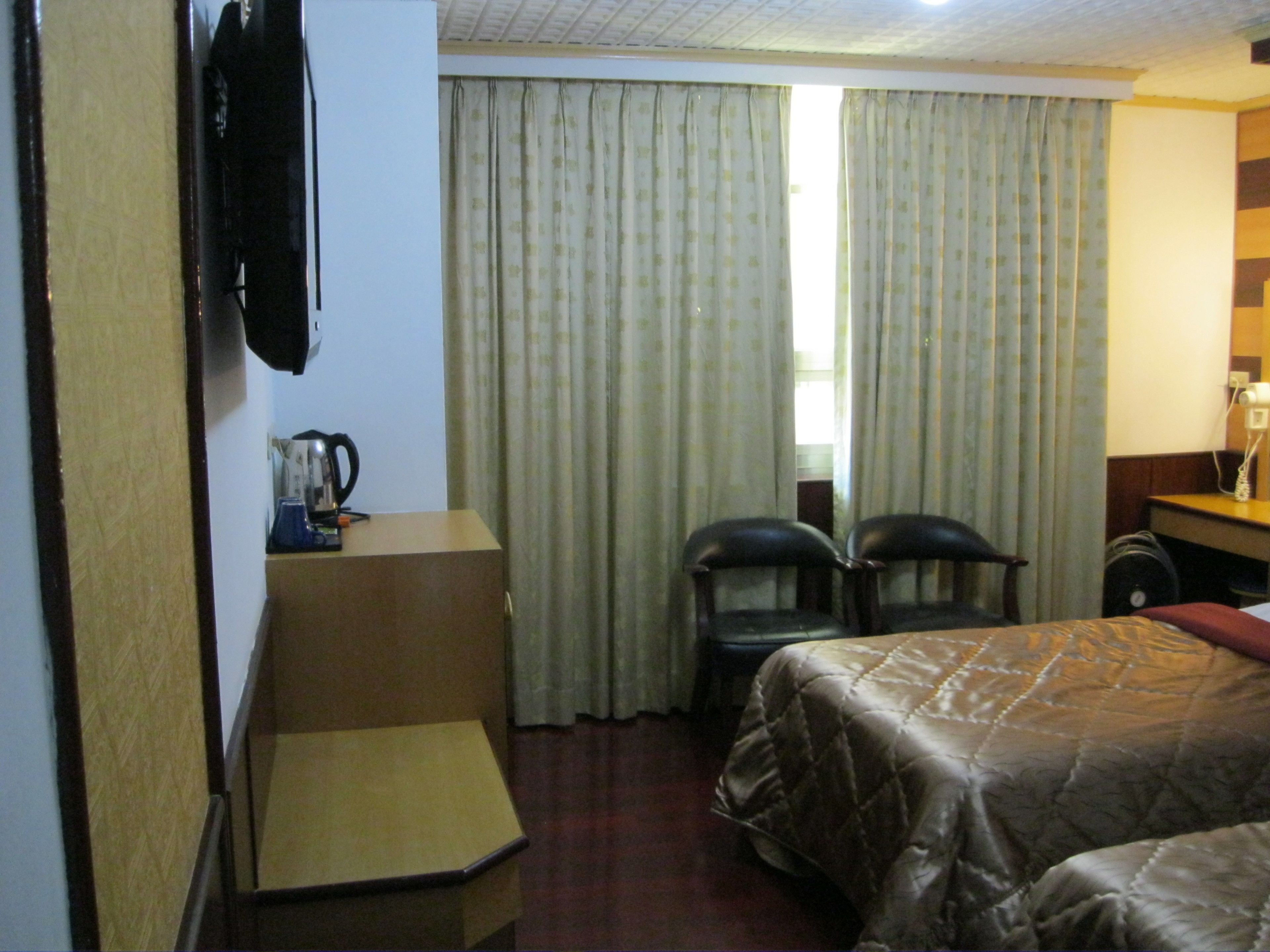 Une chambre d’hôtel comprenant un lit, deux chaises, un bureau, une télévision et une fenêtre avec des rideaux.