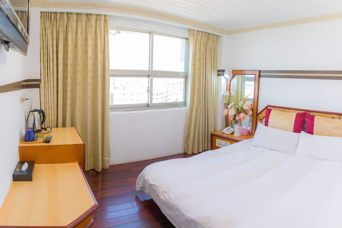 A hotel room with a bed, bedside table, a desk with a telephone, a mirror, and a window with curtains.
