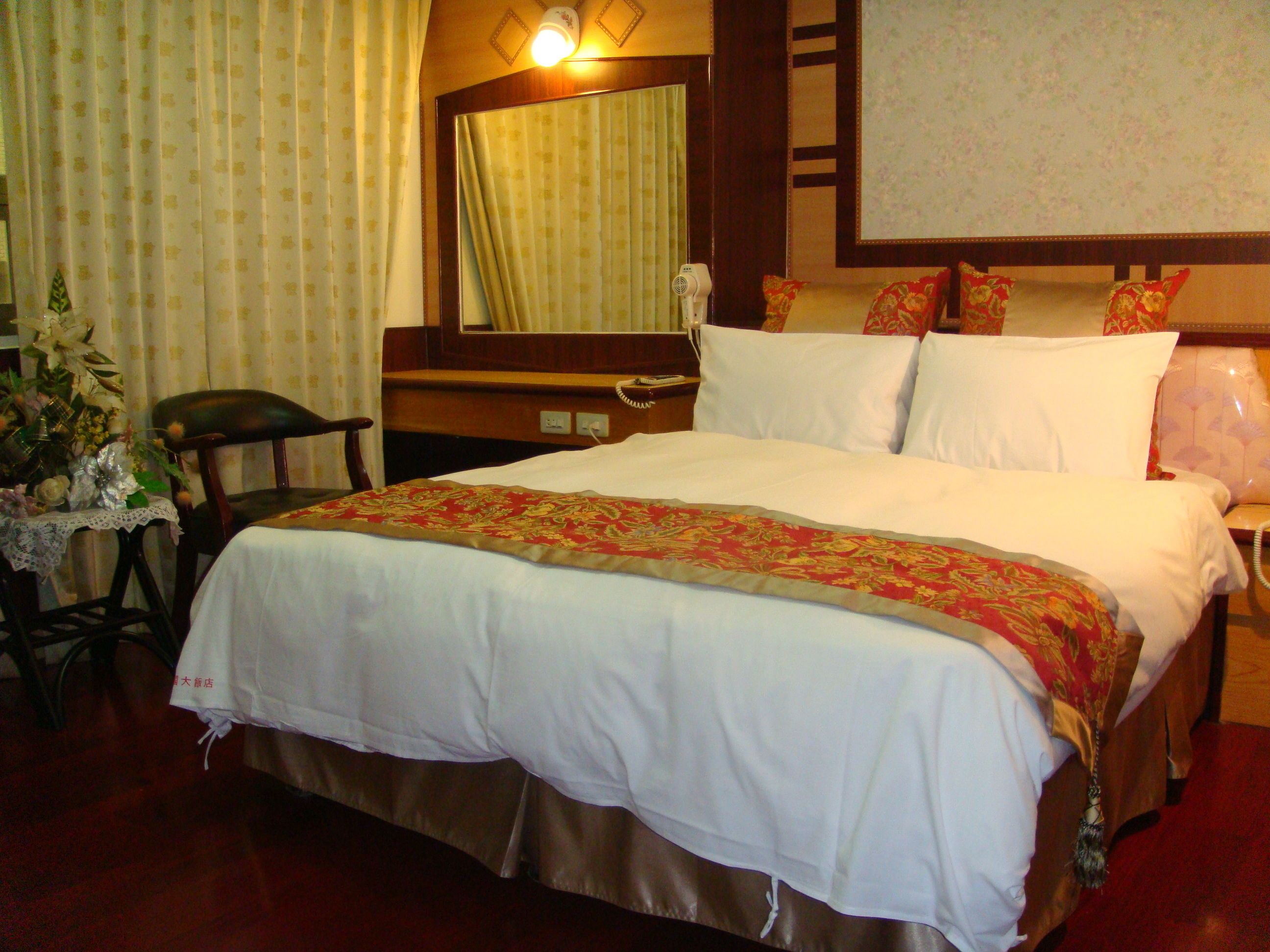 A hotel room with a bed, a chair, a table with a vase of flowers, and a mirror on the wall.