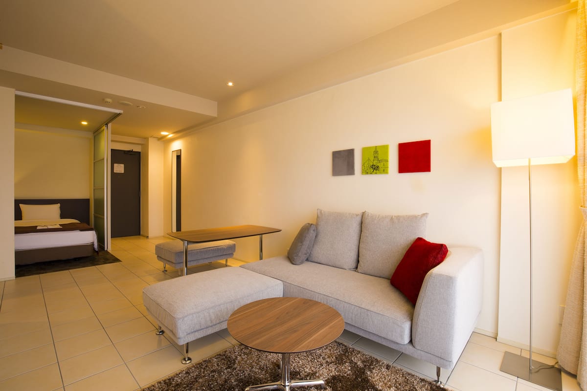 Ein modernes Wohnzimmer mit einer Couch, einem Hocker und einem kleinen runden Tisch. Im Hintergrund steht ein Bett und eine Tür, die in einen anderen Raum führt.