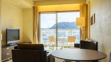A hotel room with a television, a dining table with chairs, a sofa, and a large window offering a view of a cityscape and mountains.