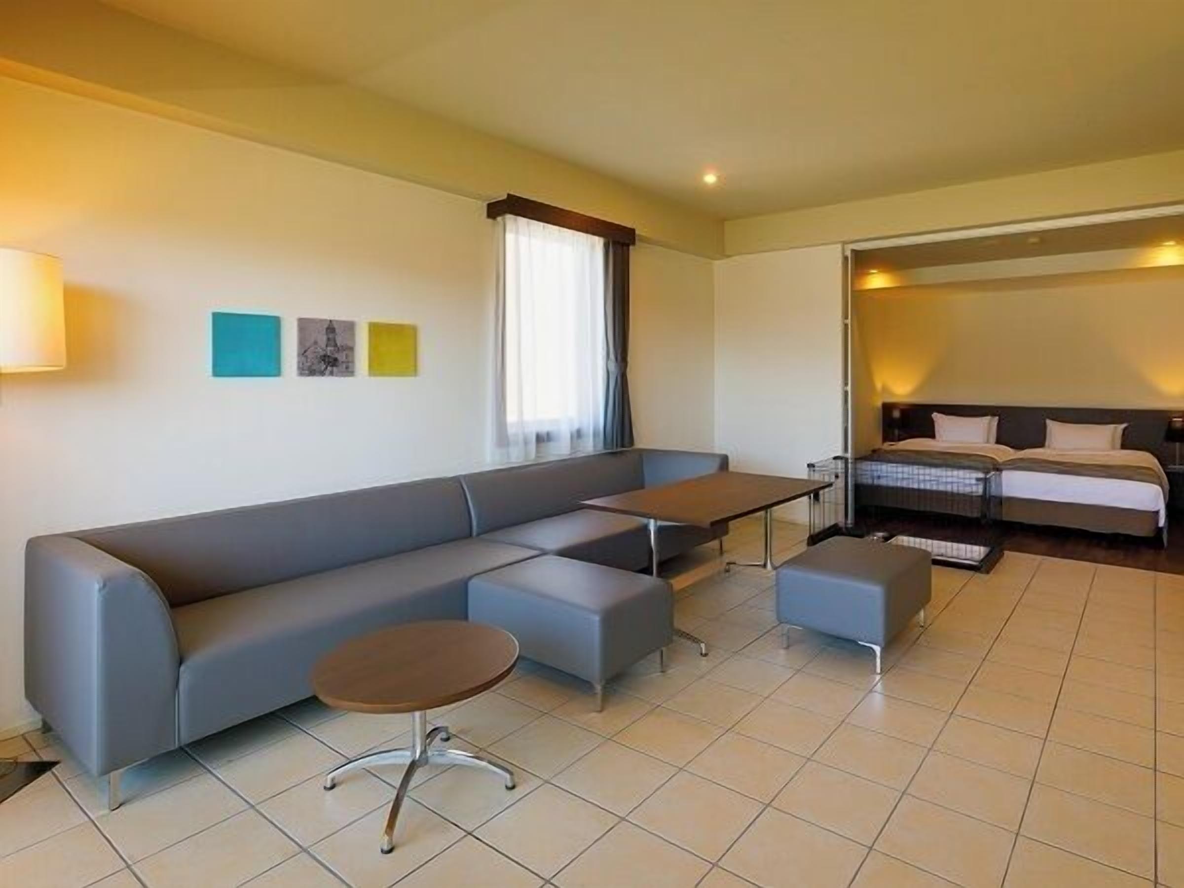 A modern hotel room with a grey sofa, a small round table, a bed with a dark headboard, and a window with curtains.