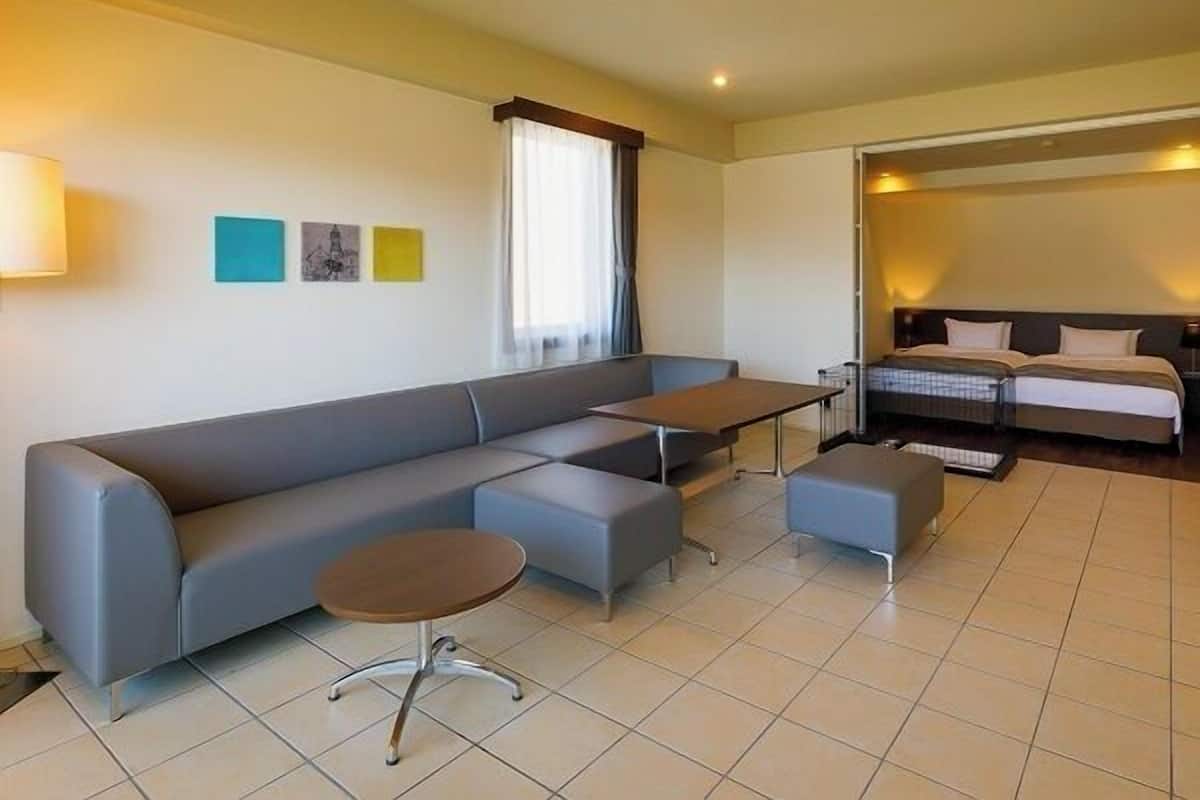 Ein modernes Hotelzimmer mit grauem Sofa, einem kleinen runden Tisch, einem Bett mit dunkler Kopfteil und einem Fenster mit Vorhängen.