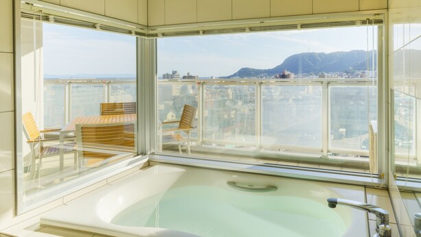 Un baño moderno con una bañera grande, cabina de ducha con mampara de vidrio y vistas a un paisaje urbano a través de ventanas amplias.