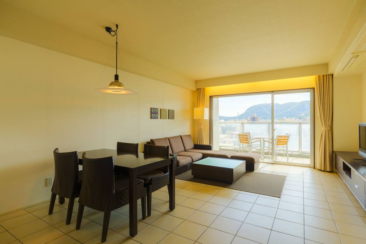 Ein modernes Wohnzimmer mit Esstisch, einer Couch und Sesseln. Ein Fernseher steht auf einem Schrank, ein großes Fenster mit Vorhängen bietet Blick auf die Stadt.