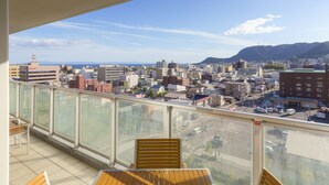 Un balcón con mobiliario de madera, baranda de cristal y vistas a un paisaje urbano con edificios y montañas.