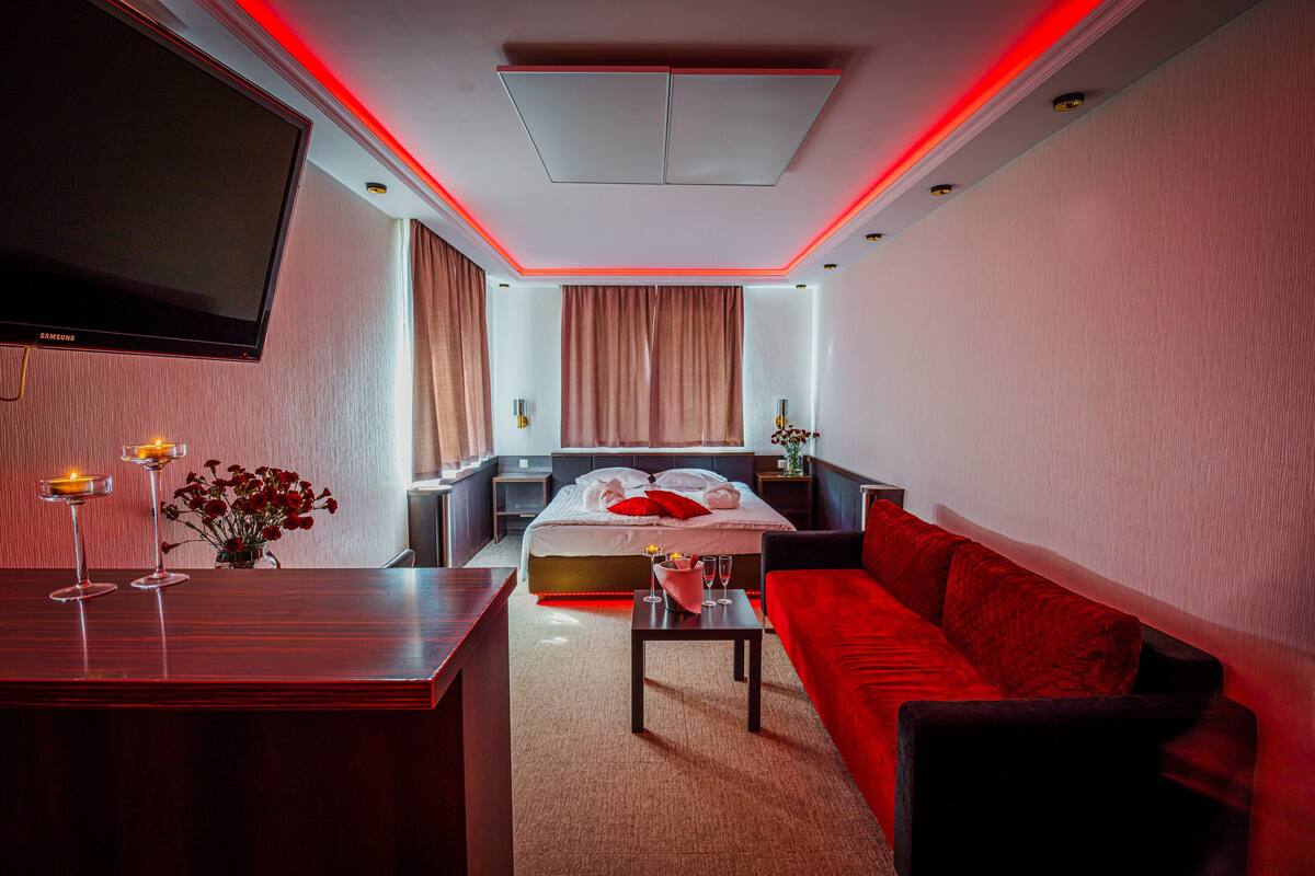 Una habitación de hotel moderna con una televisión de pantalla plana, una cama con almohadas rojas, un sofá rojo, una mesita con un florero y un escritorio con una lámpara y vasos.