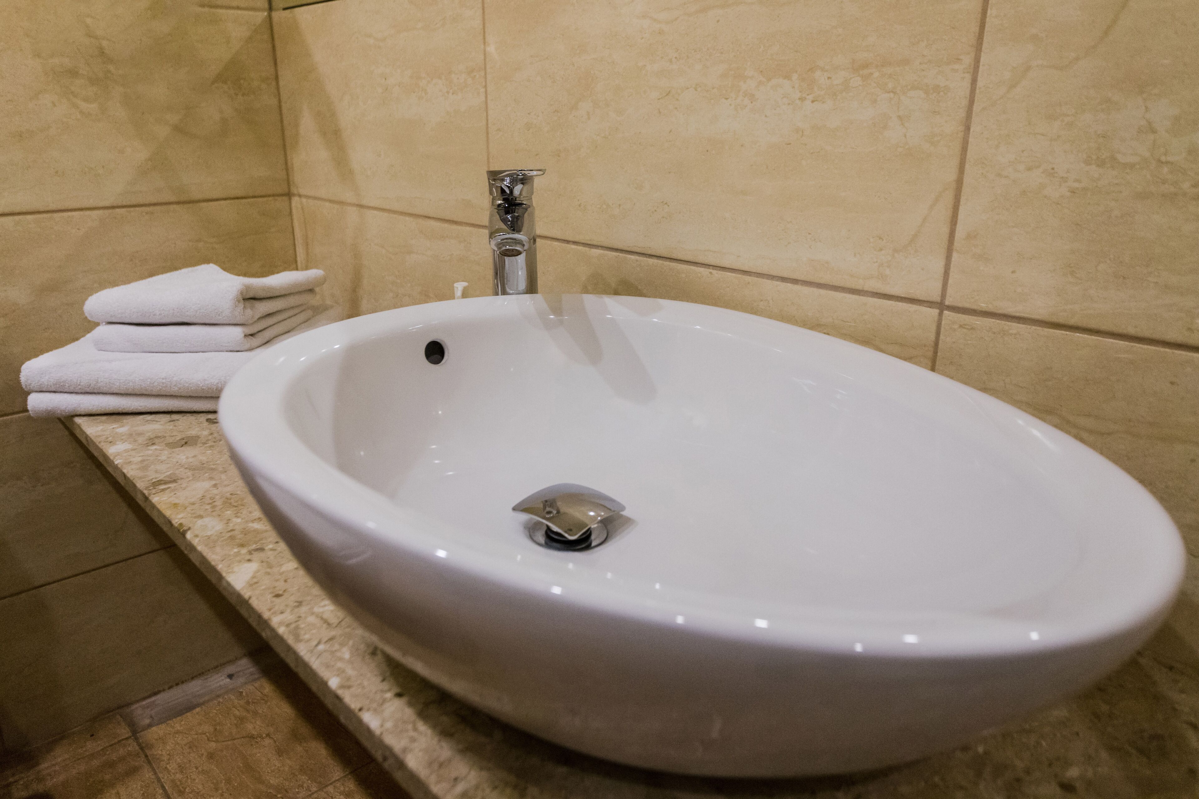 A white ceramic sink with a single faucet in a bathroom with beige tiled walls and a marble countertop.