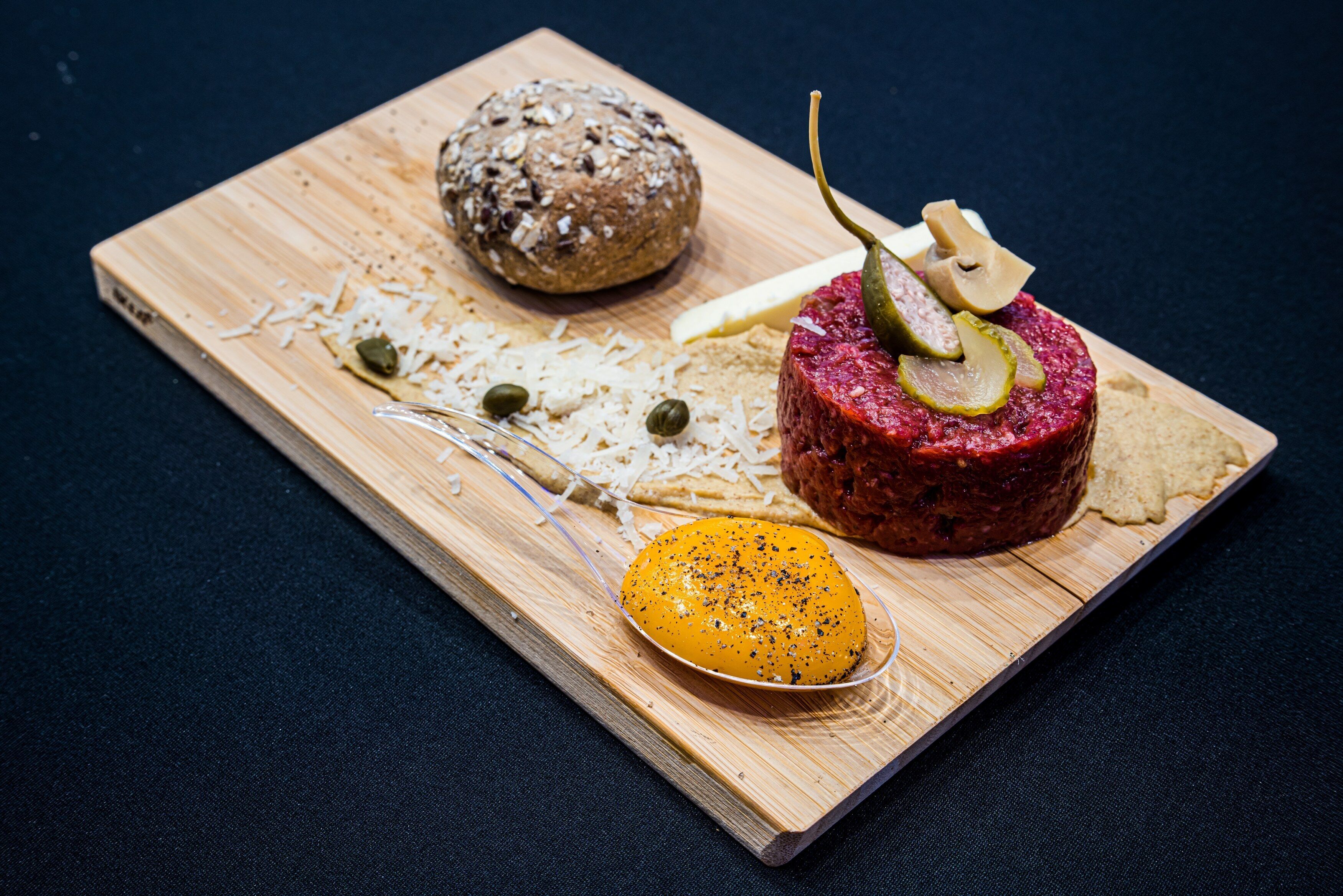 Una tabla de madera con un plato redondo de carne, una rebanada de pan y un pequeño recipiente con salsa.