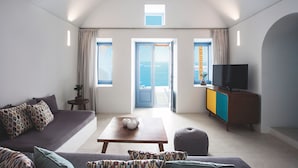 A modern living room with a sofa, a coffee table, a television, and a view of the sea through large windows. - Remezzo Villas (Santorini)