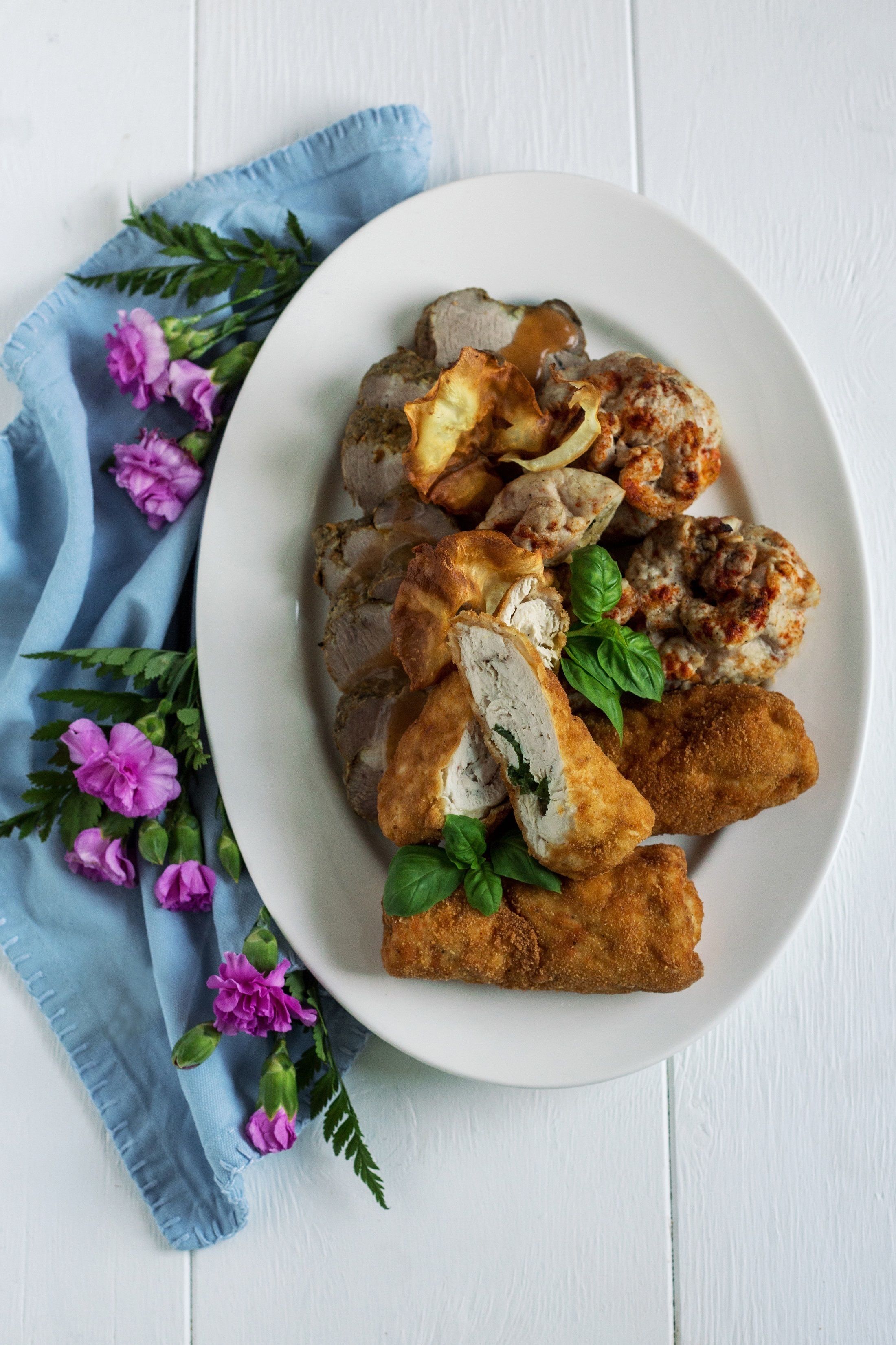 A white plate with various food items, including breaded chicken and mushrooms, on a white wooden surface with a blue cloth and purple flowers.