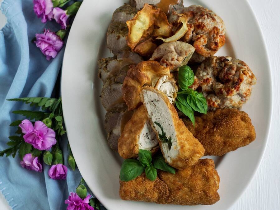 Un plato blanco con diferentes alimentos, entre ellos pollo empanizado y champiñones, sobre una superficie de madera blanca con un mantel azul y flores moradas.