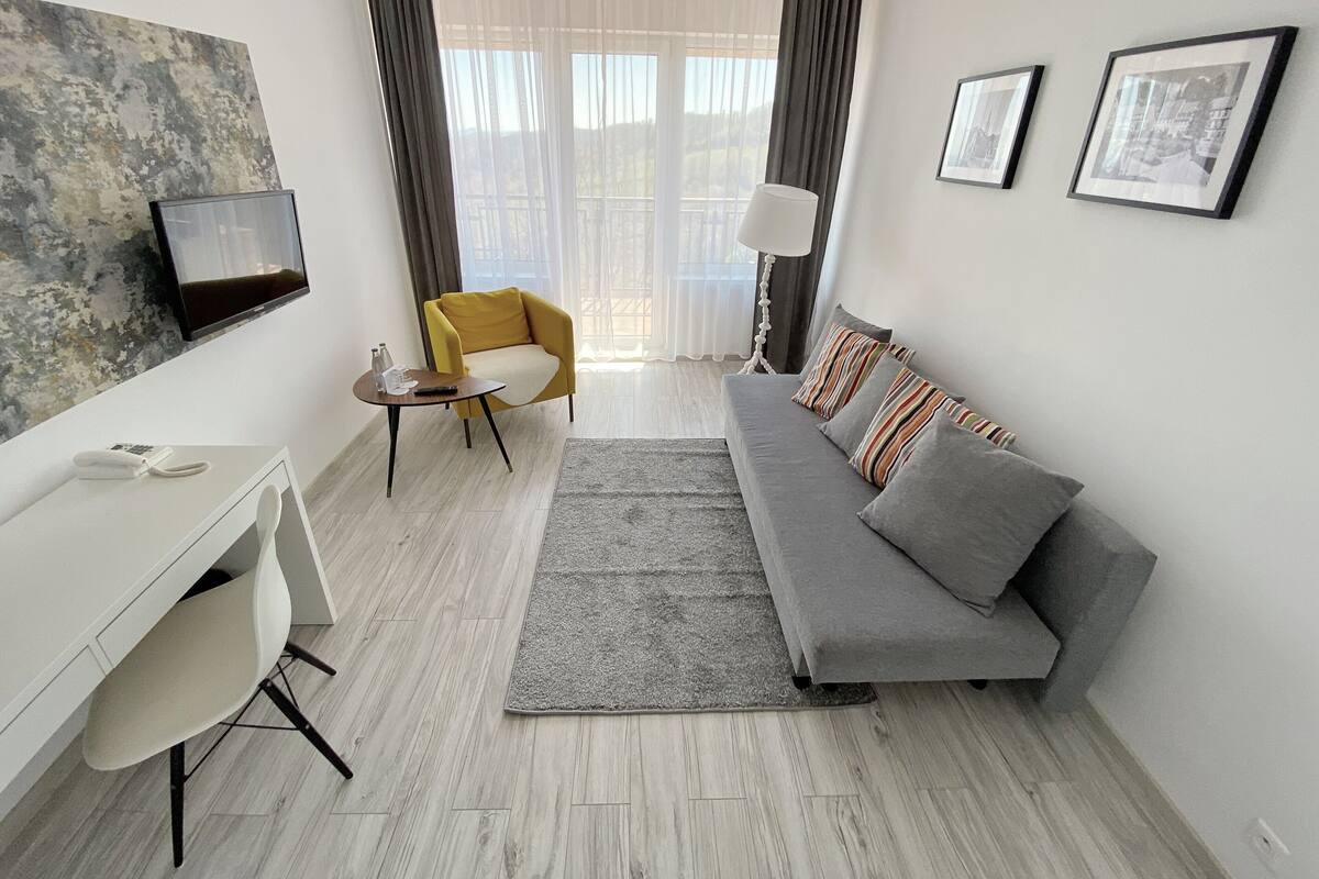 Een moderne woonkamer met een grijze bank, een gele fauteuil, een wit bureau en een flatscreen televisie aan de muur.