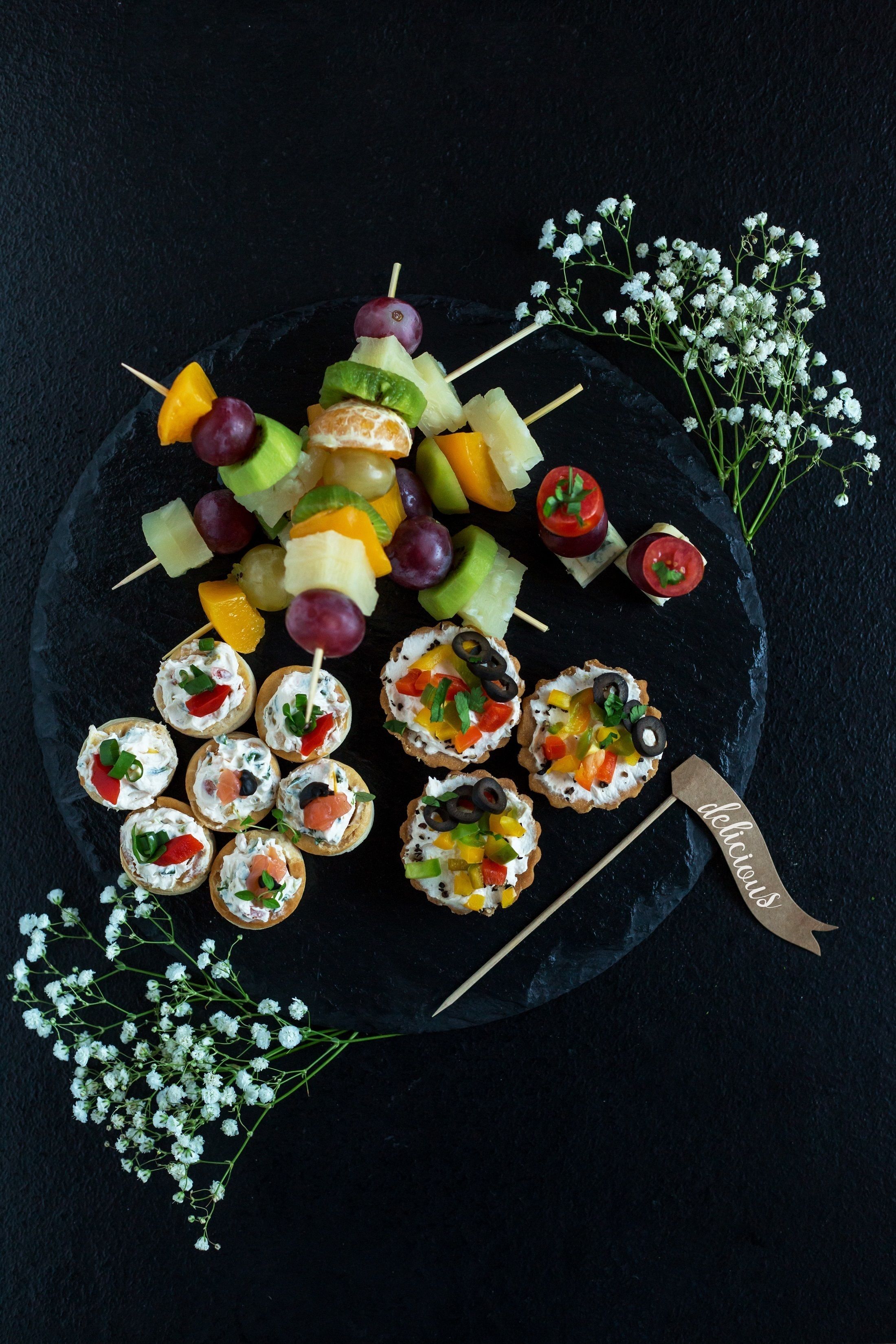 Une grande assiette remplie d’une variété de collations, notamment des brochettes de fruits et de petites bouchées, disposées sur une planche d’ardoise sombre.
