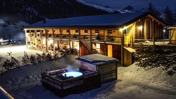 Una cabaña de madera con jacuzzi y una terraza de madera, rodeada de nieve y montañas.