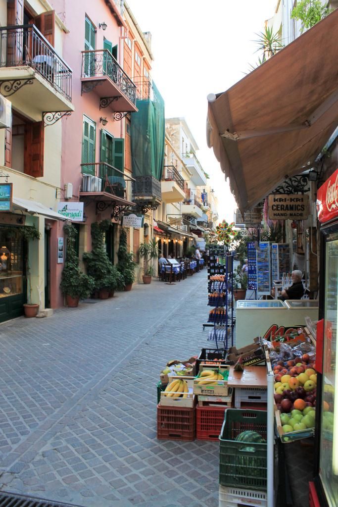 A narrow street with shops and balconies, a fruit stand, and a Coca-Cola sign.