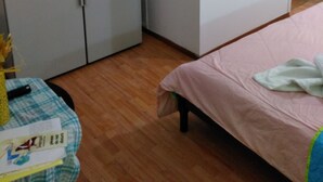 A small, compact kitchen with white cabinets, a sink, and a stove. There is a bed with a pink blanket and a small table with a placemat and a yellow object. - Smaragdi Rooms for Rent (Chania)