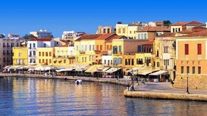 A waterfront street with colorful buildings, a canal, and outdoor seating areas. - Smaragdi Rooms for Rent (Chania)