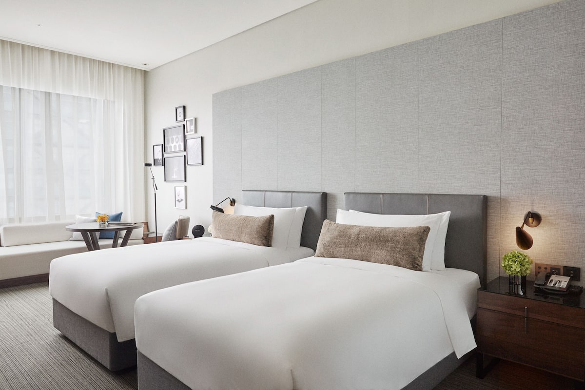 Habitación de hotel con dos camas, un sofá, una mesita, una lámpara y cuadros en la pared.