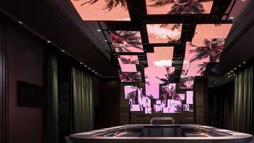Una habitación moderna con poca luz, un área de estar central, un techo con temática tropical y un amplio ventanal con cortinas.