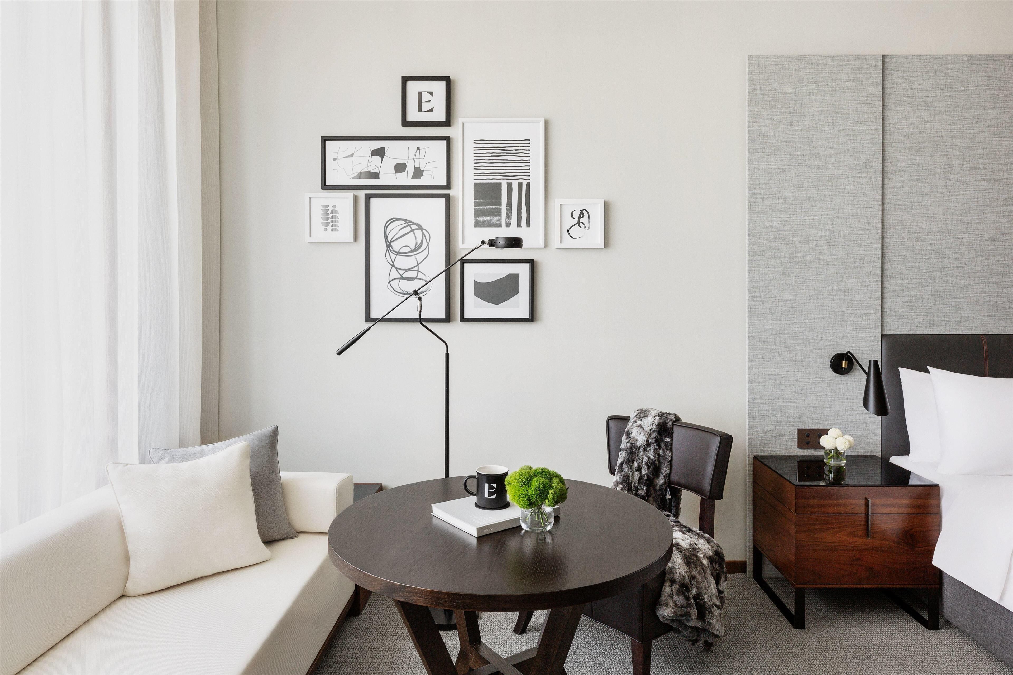 Quarto de hotel moderno com uma mesa de jantar redonda, um sofá, uma mesa de cabeceira com abajur e uma parede com obras de arte emolduradas.