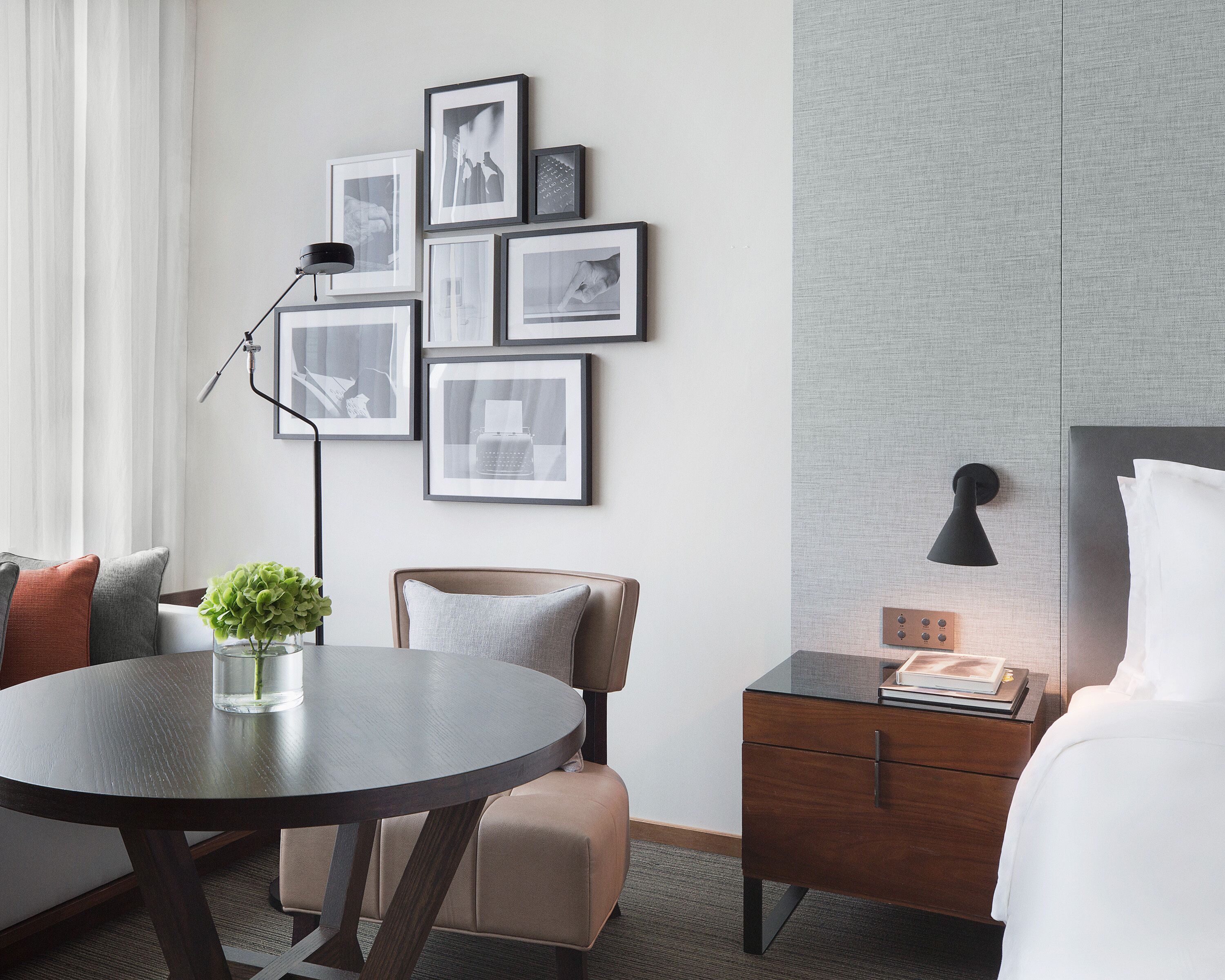 Een moderne hotelkamer met een ronde eettafel, een bank, een stoel, een nachtkastje met een lamp, en een muur versierd met ingelijste foto’s.