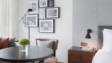 Een moderne hotelkamer met een ronde eettafel, een bank, een stoel, een nachtkastje met een lamp, en een muur versierd met ingelijste foto’s.