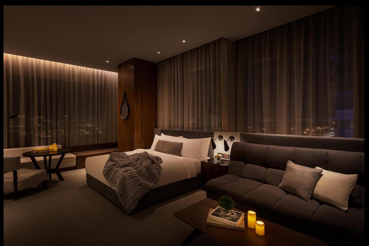 Una habitación de hotel moderna con una cama grande, un sofá, una mesita y vistas a la ciudad de noche.