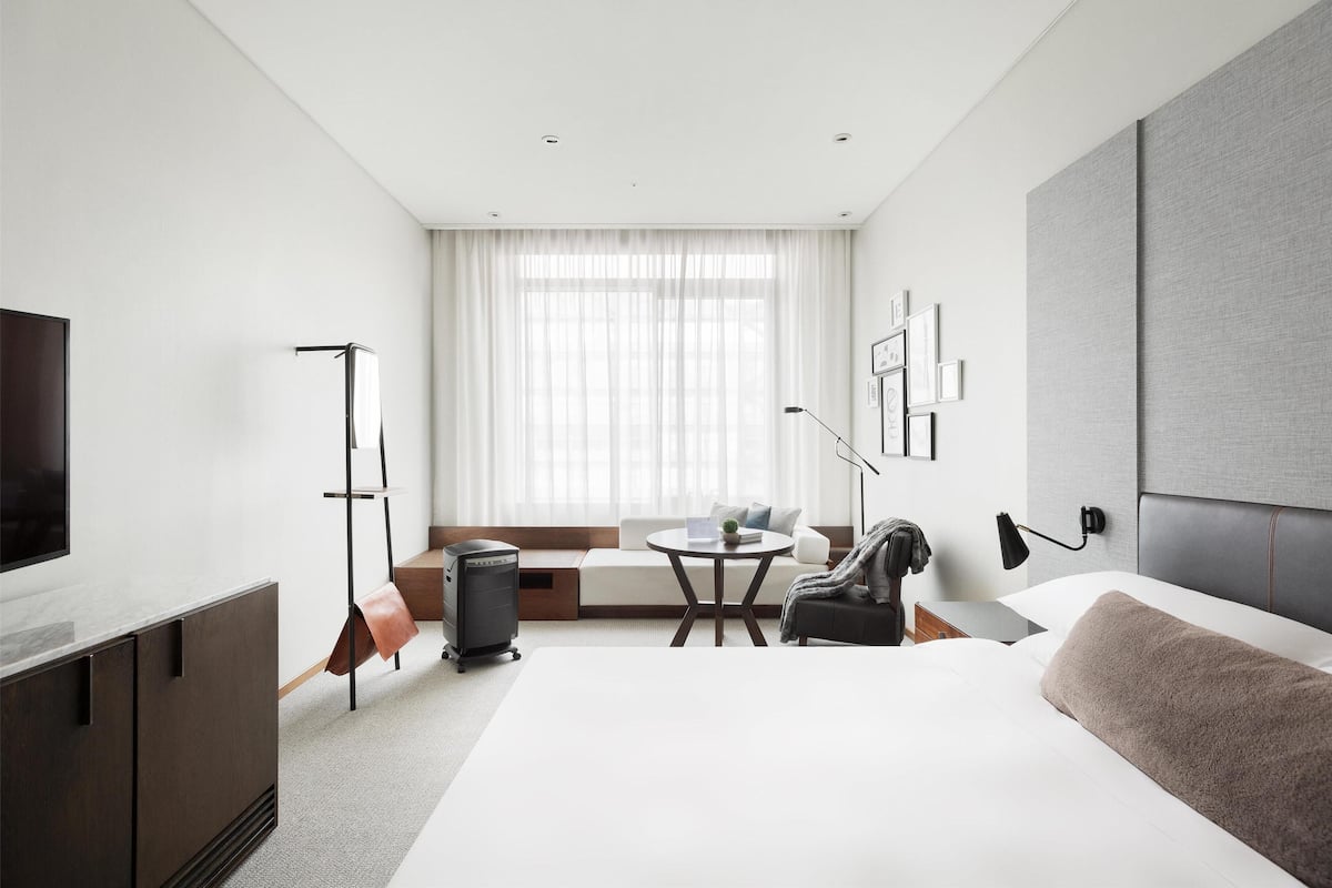 Habitación de hotel moderna con una cama grande, un escritorio, una silla, un televisor y una ventana con cortinas ligeras.