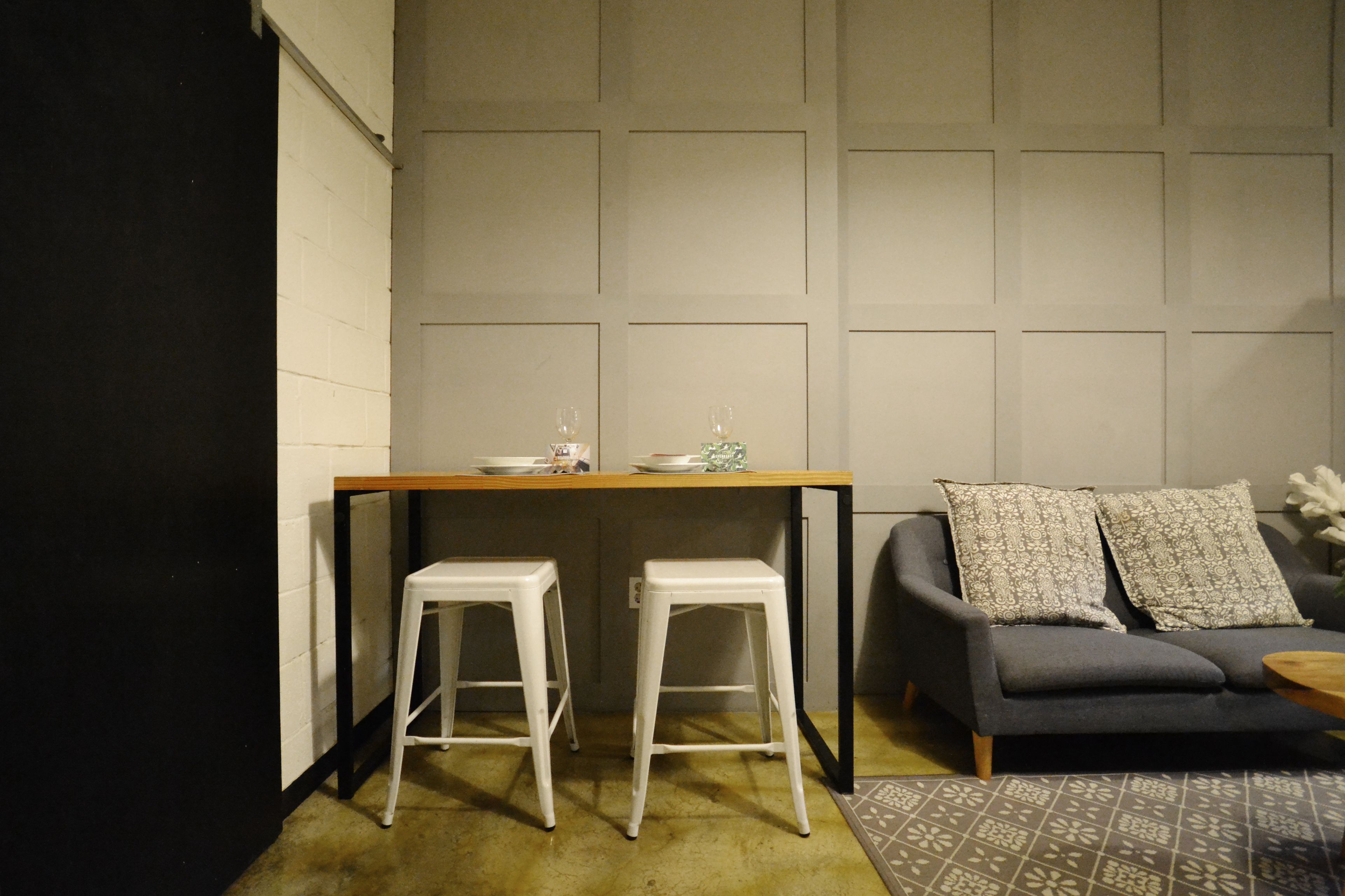 Um espaço de estar compacto com cozinha compacta, dois bancos brancos, um sofá cinza com almofadas estampadas e uma pequena mesa redonda.