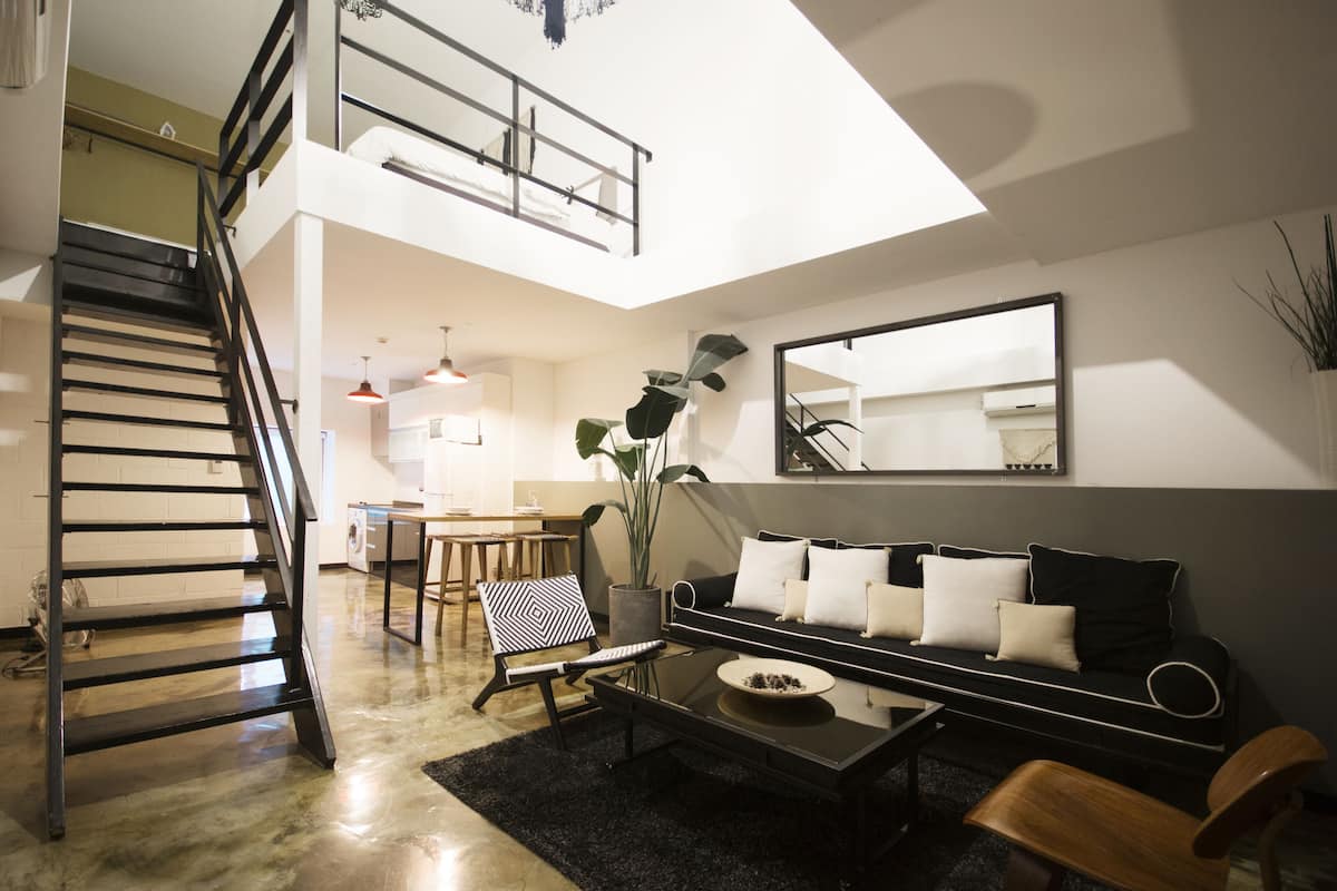 Una sala de estar moderna con un sofá negro, una mesa de centro, un sillón de estampado blanco y negro, una escalera de metal negro y un espejo grande en la pared.