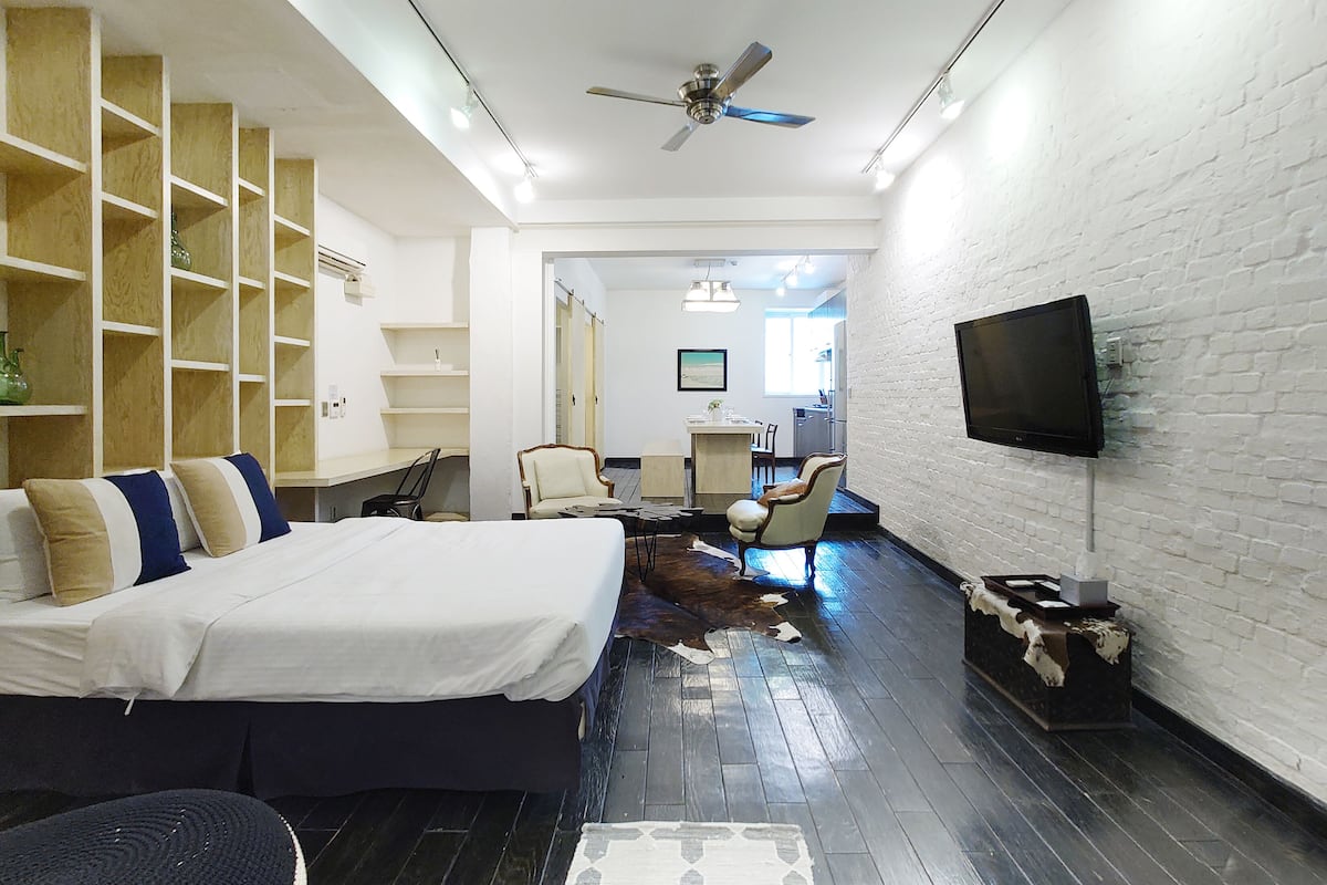 Una habitación de hotel moderna con una cama grande, un ventilador de techo, un televisor de pantalla plana montado en la pared y una estantería de madera.